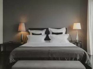 Ein modern eingerichtetes Schlafzimmer mit einem großen Doppelbett in der Mitte. Das Bett ist mit einer grauen Bettdecke und mehreren weißen Kissen sowie einem schwarzen Zierkissen ausgestattet. Zu beiden Seiten des Bettes stehen Nachttische, auf denen jeweils eine Tischlampe mit silberfarbenem Fuß und weißem Schirm das Zimmer in ein warmes Licht taucht. Die Wand hinter dem Bett ist in einem dunklen Grauton gehalten.