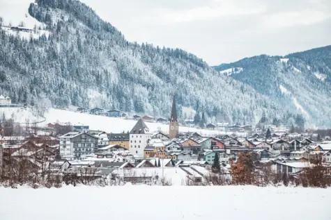 Ein in Schnee getauchtes Dorf mit vielen bunten Gebäuden. Im Hintergrund befinden sich Tannenbewachsene Berge und weite, schneebedeckte Felder. 