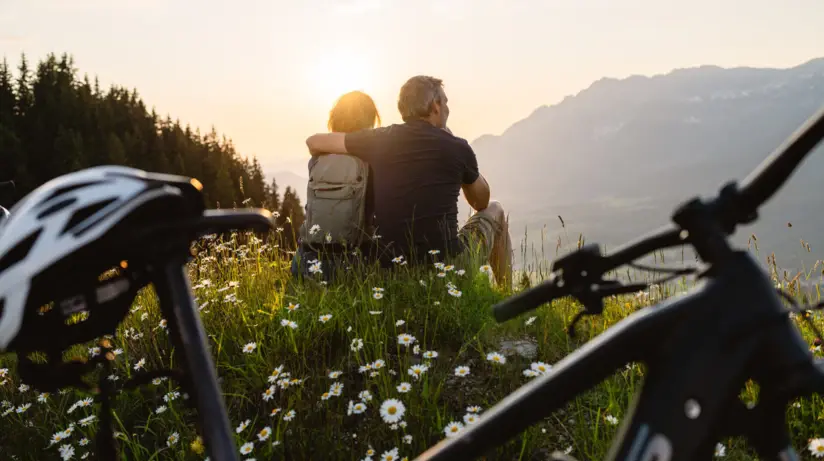 Ein Paar sitzt Arm in Arm auf einer blumigen Wiese. Direkt vor der Kamera steht eines ihrer Fahrräder und im Hintergrund scheint die warme Sommersonne auf hohe Berge. 