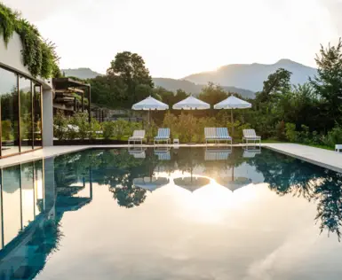 Swimmingpool im Freien umgeben von Stühlen und Sonnenschirmen mit Bäumen und einem klaren Himmel mit untergehender Sonne im Hintergrund.