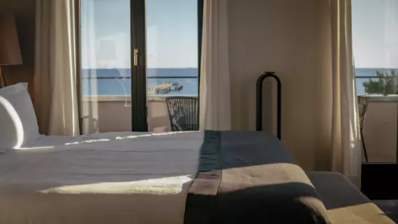 Ein sonnendurchflutetes Hotelzimmer mit bodentiefen Fenstern. Von der Seite ist ein großes, weißes Bett mit dunkler Tagesdecke zu sehen und im Hintergrund die offene See.. 