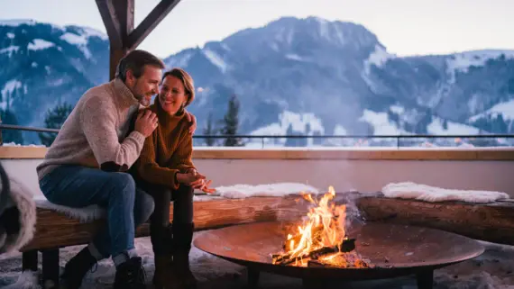 Ein Paar sitzt gemütlich an einer Feuerstelle auf einer Terrasse, umgeben von einer verschneiten Berglandschaft in der Dämmerung. 