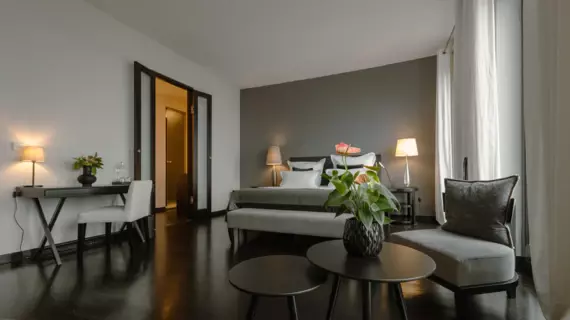  Im Vordergrund des Hotelzimmers stehen ein runder Couchtisch und ein gemütliches Sofa, auf dem ein einzelnes, samtiges Zierkissen liegt. Im Mittelpunkt des Raumes befindet sich ein großes Bett, dekoriert mit weißen und grauen Kissen, die einen eleganten Kontrast zur dunklen Wand im Hintergrund bilden.