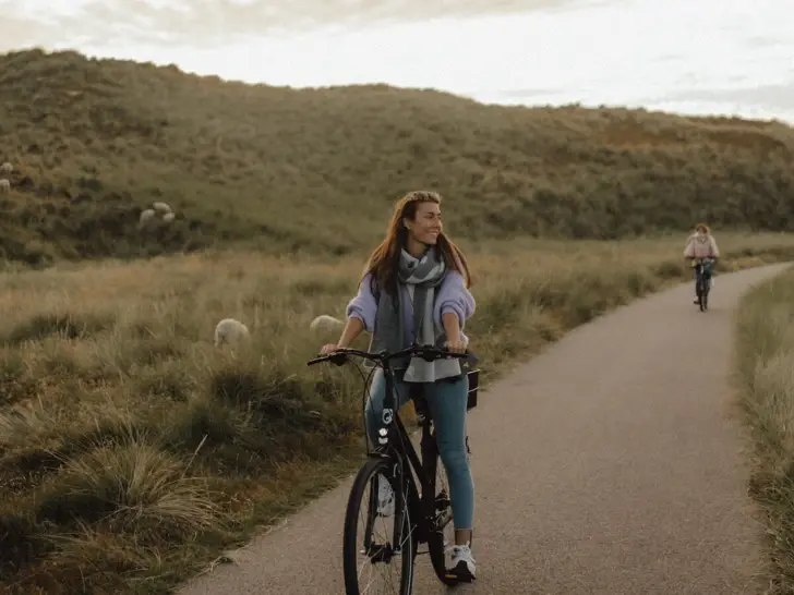 Eine Frau fährt lächelnd auf einem Fahrrad einen asphaltierten Weg entlang. Um sie herum sind grüne Dünen auf denen vereinzelt Schafe grasen und die Sonne scheint durch die Wolkendecke. 