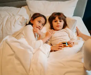Zwei Kinder liegen in einem Hotelbett und werden von einer Frau ins Bett gebracht. Das Mädchen auf der rechten Seite hält ein Spielzeugauto in den Händen.