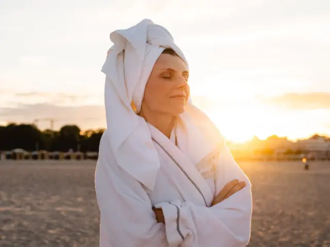 Eine Frau steht in einen Bademantel gewickelt mit geschlossenen Augen am Strand und genießt die aufgehende Sonne.