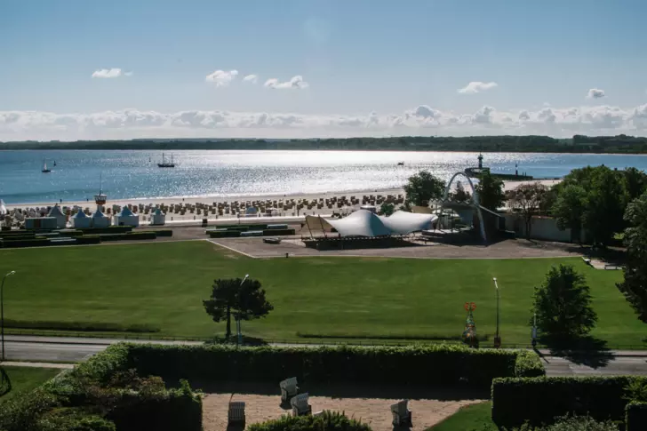 Ein Hotelaußenbereich ist durch eine waagerecht verlaufende Straße von einer dahinter liegenden Wiese und dem Strand mit Strandkörben getrennt. Das Wasser mit Booten glitzert im Licht der Sonne.