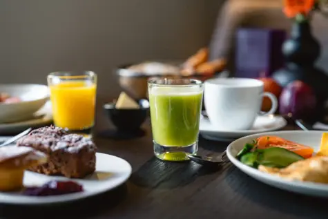 Multiple different drinks like a smoothie and orange juice on a breakfast table with bread and other dishes. 