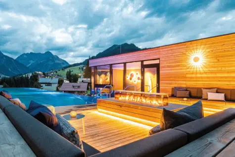 Modern gestalteter Außenbereich des SPA-ROSA im A-ROSA Ifen mit beheiztem Infinity-Pool, Feuerstelle, gemütlichen Loungemöbeln und Blick auf die Berge des Kleinwalsertals bei Abendstimmung.
