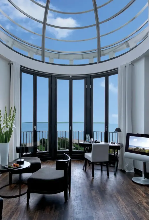 A room with a glass domed roof. In front of three floor-to-ceiling windows with dark frames, there is a desk and a round side table, each with an upholstered chair on a dark parquet floor.
