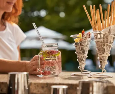 Eine Frau sitzt an einer Bar und hält ein Glas Limonade in ihrer Hand. Neben ihr stehen zwei Snacks.