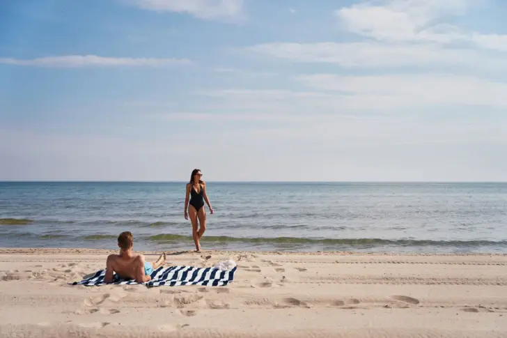 Ein Mann liegt an einem sonnigen Tag auf einem blau weiß gestreiften Handtuch am Strand und eine Frau in schwarzem Badeanzug geht vor Meer auf ihn zu. 