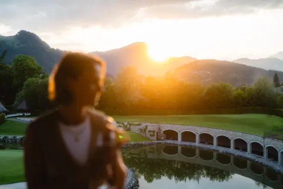 Frau steht an einem Teich mit einer Brücke und Bergen im Hintergrund.