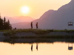 Zwei Wanderer genießen einen Spaziergang bei Sonnenuntergang entlang eines stillen Sees, umgeben von einer malerischen Berglandschaft, deren Silhouetten im Wasser reflektieren.