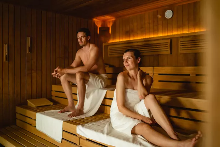 Ein Mann und eine Frau entspannen sich in einer Sauna; der Mann sitzt auf einer höheren Bank, während die Frau auf einer niedrigeren Bank sitzt, beide eingehüllt in Handtücher und genießen die Wärme um sie herum.