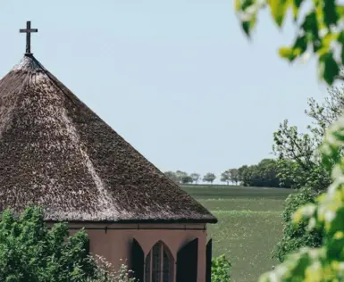 Eine kleine runde Kapelle mit Strohdach steht auf einer grünen Wiese.