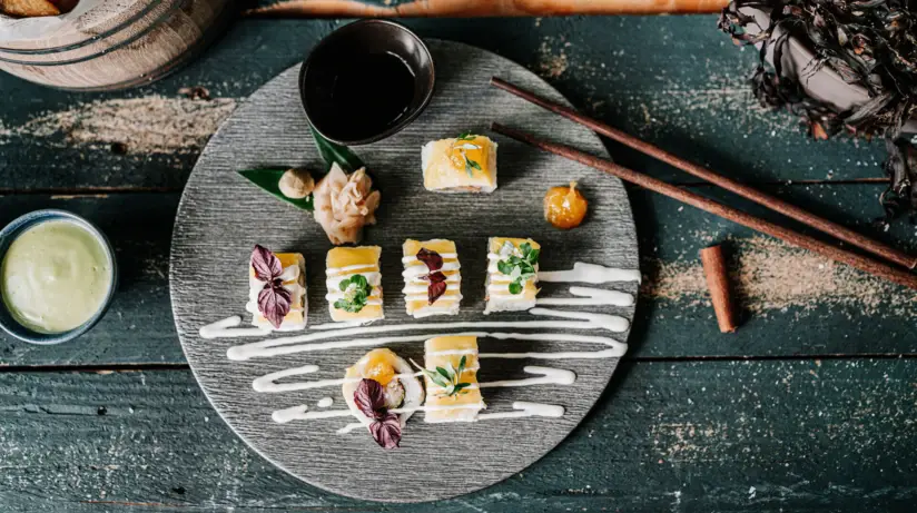 Kunstvoll angerichtete Sushi-Variationen auf einer runden Schieferplatte, serviert auf einem rustikalen Holztisch. Die Sushi-Stücke sind garniert mit frischen Kräutern und essbaren Blüten, begleitet von Sojasauce, Wasabi und eingelegtem Ingwer. Im Hintergrund sind verschwommen weitere Tischbestandteile wie Stäbchen und ein Schälchen mit grüner Sauce zu erkennen. Die Komposition ist sorgfältig arrangiert und bietet eine visuell ansprechende Darstellung der japanischen Küche.