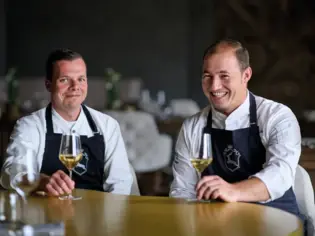 Two men in aprons sitting at a table, smiling and holding wine glasses.