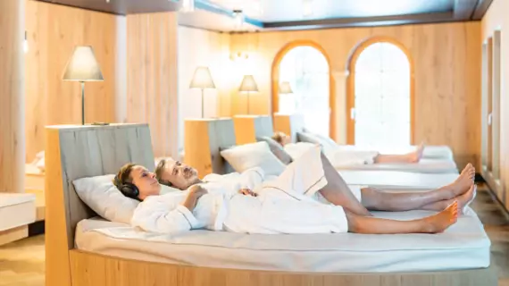Ein entspanntes Paar in weißen Bademänteln liegt bequem auf einer Liege in einem hellen, holzgetäfelten Raum, während sie in einem ruhigen Spa-Bereich Erholung genießen.