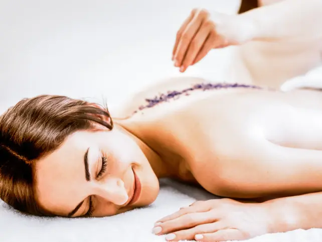 Eine Frau liegt auf ihrem Bauch mit freiem Rücken auf einem weißen, flauschiegen Handtuch. Sie lächelt und entlang ihrer Wirbelsäule streut eine Hand Lavendel. 