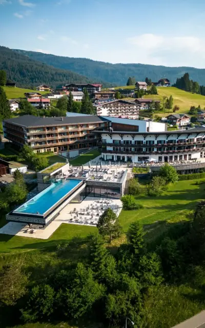 A building with an infinity pool in the middle of a mountain village, surrounded by trees and green spaces.