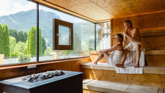 Ein Paar sitzt lächelnd in einer Sauna mit großer Glaswand, die auf eine grüne und bergige Landschaft blicken lässt. 