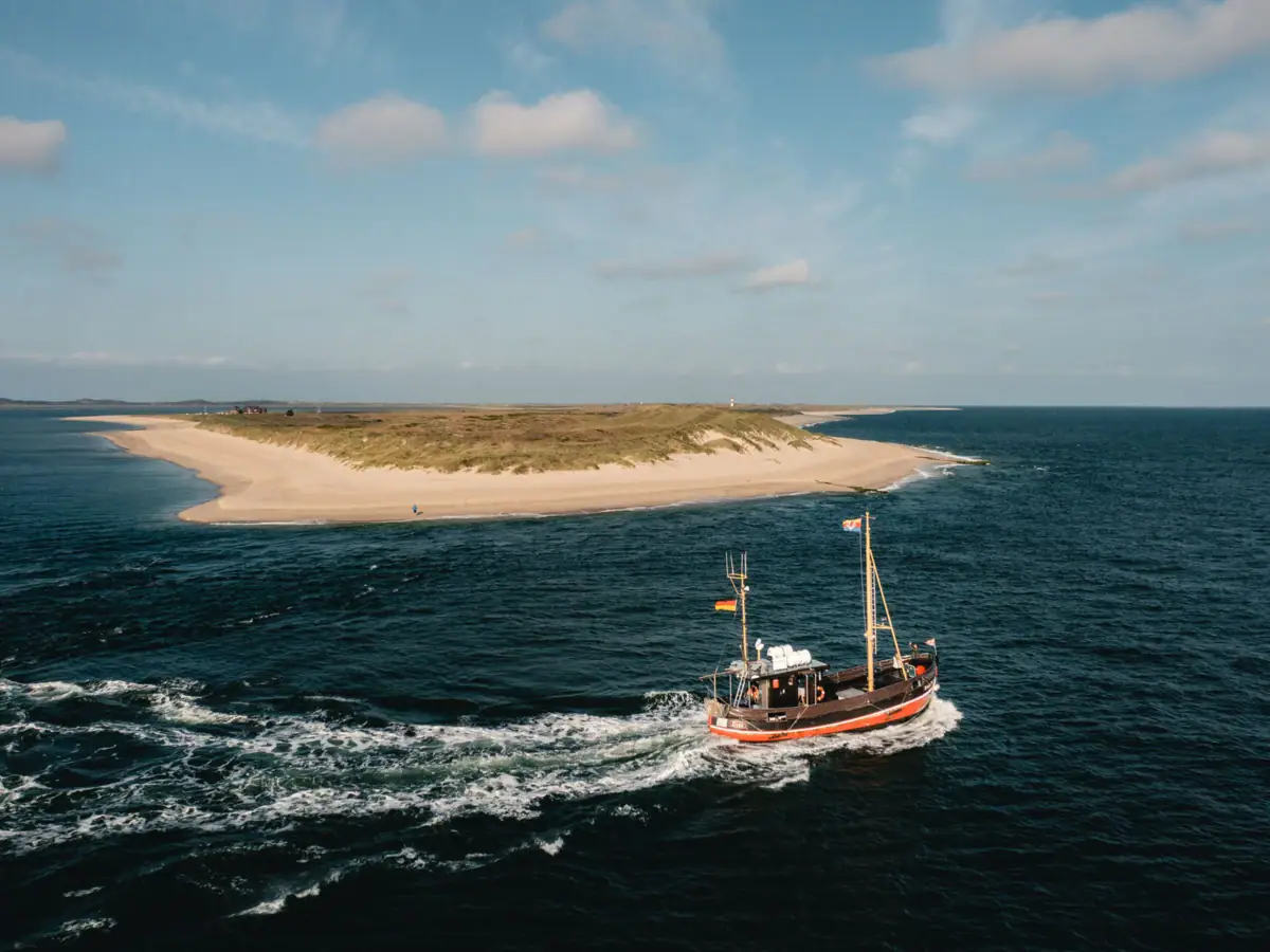 Bootstour Sylt Luftaufnahme eines traditionellen Fischerboots auf offener See nahe einer malerischen Sandbank unter einem klaren blauen Himmel. Die Szene strahlt Ruhe und Abgeschiedenheit aus, während das Boot sachte Wellen auf dem Wasser hinterlässt und sich seinen Weg durch das Meer bahnt. Im Hintergrund ist die weite, unberührte Landschaft zu erkennen, die die Naturschönheit der Küstenregion hervorhebt.