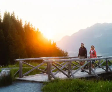 Zwei Frauen wandern in einem grasbewachsenen Feld neben einem See, umgeben von Bergen und Bäumen.