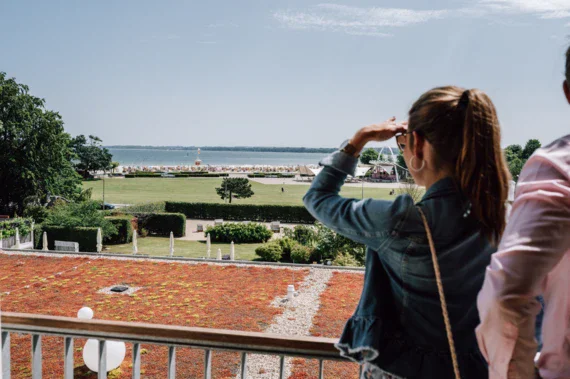 Ein Bild zeigt zwei Personen, die von einer Balkonbrüstung aus auf eine Strandpromenade und die Ostsee blicken. Die Frau im Vordergrund, mit hochgesteckten Haaren und einer Jeansjacke, hält sich die Hand über die Augen, um besser in die Ferne sehen zu können. 