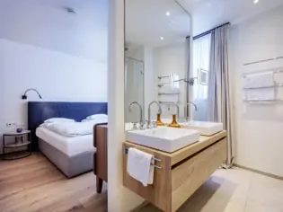 Ein Schlafbereich, der durch eine kleine Wand in der Mitte zum Badezimmer offen gestaltet ist. An der kleinen Wand ist ein Waschtisch aus Holz mit zwei Waschbecken angebracht und im Hintergrund ist ein blaues Polsterbett mit weißer Bettwäsche zu sehen.