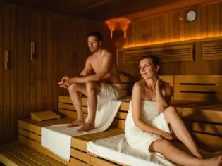 Ein Mann und eine Frau sitzen in Handtücher gewickelt in einer warm beleuchteten Holzsauna.  