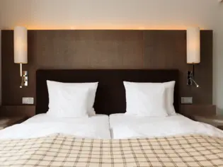Ein Bett in einem Hotelzimmer frontal fotografiert mit vier weißen Kissen und einer karierten Tagesdecke am Fußende. An beiden Seiten des Bettes stehen zwei Nachttische und es hängen jeweils eine Lampe an der Wand.