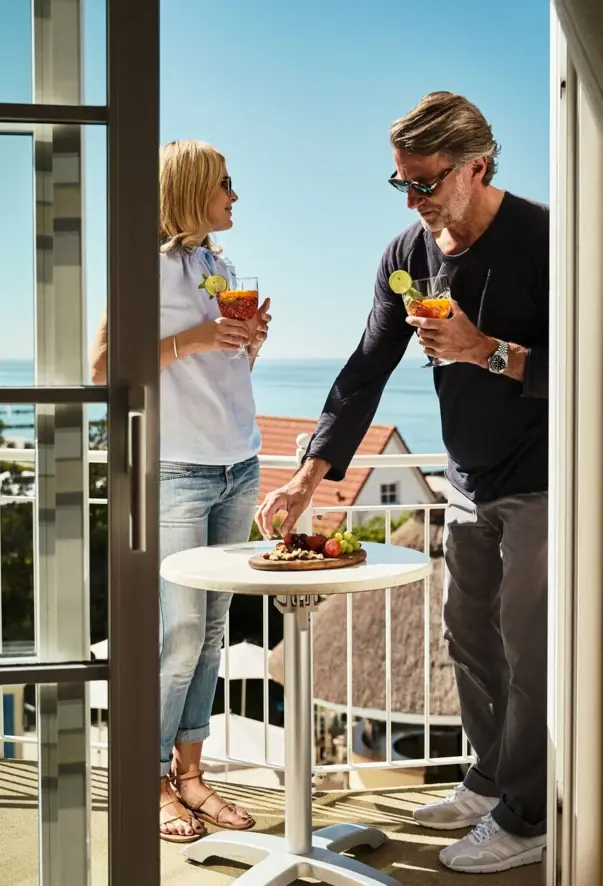 Ein Mann und eine Frau stehen mit Getränken auf einem Balkon mit Meerblick.