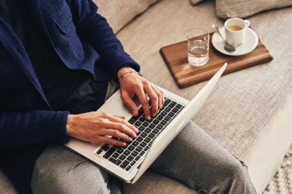 Person sitzt auf einem Sofa und benutzt einen Laptop, neben ihr steht eine Kaffeetasse.