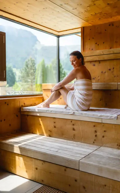 Eine Frau sitzt lächelnd in ein Handtuch gewickelt in einer Holzsauna mit großem Fenster, welches den Blick in die grün bewachsene Natur frei gibt. 