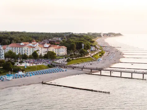 Panoramaaufnahme der Ostseeküste mit einem charmanten Strandhotel im klassischen Bäderstil, umgeben von grüner Natur und einer lebhaften Strandpromenade. Der breite Sandstrand ist gesäumt von Strandkörben und einer langen Seebrücke, die ins ruhige Meer hinausragt.