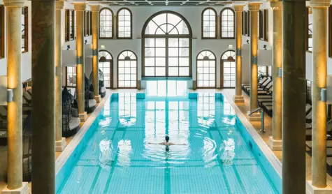 Luxuriöser Innenpool im A-ROSA Hotel Kitzbühel mit klarem, blauem Wasser, umgeben von eleganten Säulen und Bogenfenstern, die Tageslicht einlassen. Eine Person schwimmt entspannt in der Mitte des Pools, umgeben von einer ruhigen und erholsamen Atmosphäre.