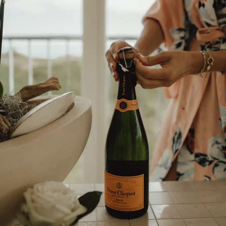 Hände einer Person, die gerade dabei ist, eine Flasche Veuve Clicquot Champagner zu öffnen. Die Flasche steht auf  der hellen Kante einer Badewanne. Im Vordergrund ist ein Zweig mit weißen Orchideenblüten und eine einzelne weiße Rose zu sehen.