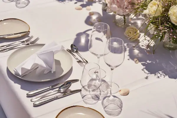 The picture shows a festively laid table outdoors, artfully arranged under bright sunlight. On the table are white porcelain plates, finely folded cloth napkins and a selection of neatly polished cutlery, which gleam elegantly in the sun. Several glasses of different sizes are set out for water and wine. The table setting also includes delicate floral arrangements of roses and wildflowers in small vases, adding natural beauty and a festive atmosphere. The scene exudes elegance and an inviting atmosphere, ideal for a special event or an elegant outdoor meal.