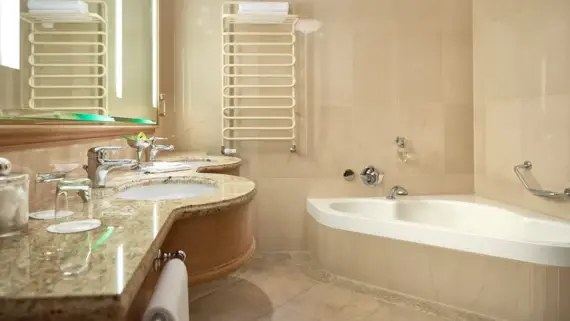 A bright and inviting bathroom that offers a blend of functionality and elegance. The room is decorated in neutral beige and cream tones, creating a warm and relaxing atmosphere. In the center of the room is a large, white bathtub that invites you to relax. Next to the bath is a large washbasin with a marble countertop and wooden elements that emphasize the natural theme of the room. A large mirror hangs above the washbasin, visually enlarging the room. There is a modern, white towel warmer on the wall for added convenience.