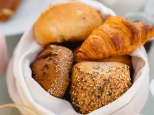 Ein Brötchenkorb von oben fotografiert. In dem hellen Korb befindet sich ein Croissant, zwei Körnerbrötchen und ein helles Brötchen. 