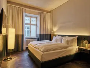 Ein Hotelzimmer mir einem großem Bett mit weißer Bettwäsche und im Hintergrund ist ein Fenster zu sehen.