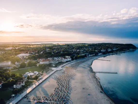 Eine Luftansicht des Küstenbereichs von Travemünde. Das Wasser leuchtet in warmem Blau, der Strand zieht sich entlang der Küste und viele Strandkörbe stehen hierauf verteilt und die Stadt liegt ruhig zwischen vielem Grün im Hintergrund. 
