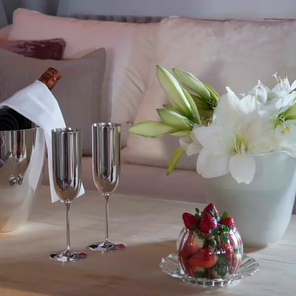 On a coffee table is a bucket of champagne, two champagne glasses, a bowl of strawberries and a vase of flowers.