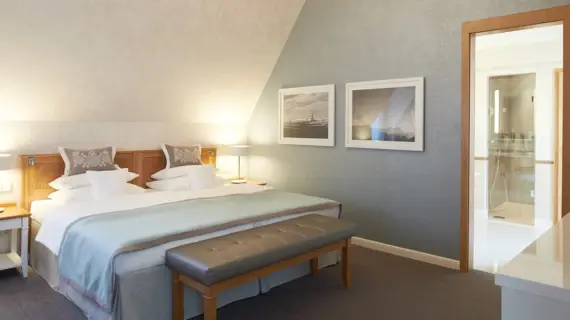 A quiet bedroom in a suite under a sloping roof. A comfortable bed with a light blue bedspread and decorative cushions with a subtle pattern takes center stage. At the foot of the bed, an upholstered bench invites you to linger. Two framed black and white photos of maritime scenes hang on the wall. An open door provides a view into an elegant bathroom with a glass shower cubicle.