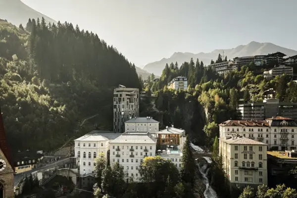 Einige Gebäude stehen in einer Berglandschaft mit vielen Tannen und einem Wasserfall, der zwischen den Häusern den Berg hinter fließt.