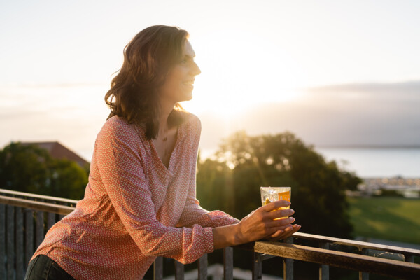 Eine Frau steht lächelnd mit einem Getränk in ihrer Hand und auf ein Geländer gestützt auf einer Terrasse. Sie schaut in die Ferne und im Hintergrund ist weite Natur und das angrenzende Meer zu sehen.