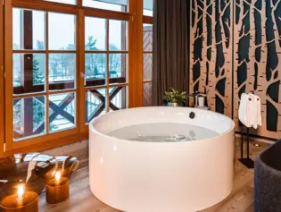 A free-standing, round bathtub stands in front of a window with a view of a snowy winter landscape. Next to the bathtub are two lamps, a plant and a towel rail with two towels.