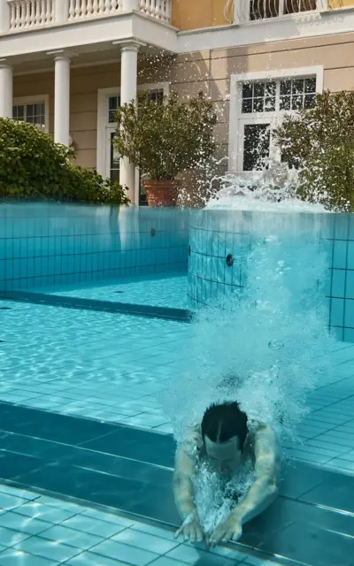 Mann schwimmt unter Wasser in einem Außenpool vor einem gelben Gebäude mit weißen Akzepten im Stil der Bäderarchitektur.