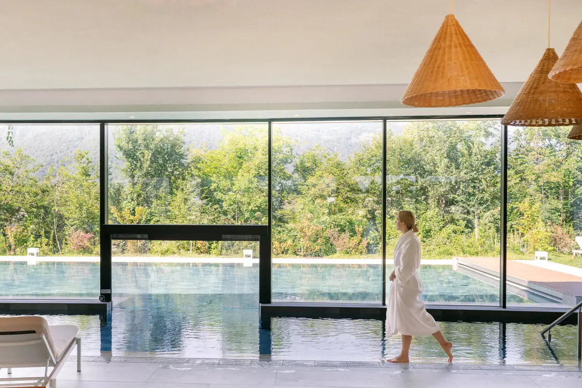  Frau im Bademantel spaziert am Innenpool des A-ROSA Gardasee entlang, mit Blick auf den Außenpool und die umliegende Natur.